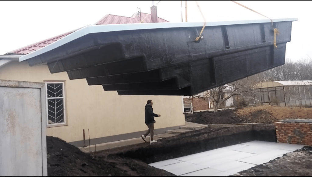 Большая черная чаша композитного бассейна Эклипс 7.6м поднята краном над подготовленным котлованом на объекте в Днепре.