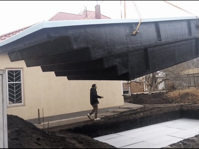 Большая черная чаша композитного бассейна Эклипс 7.6м поднята краном над подготовленным котлованом на объекте в Днепре.