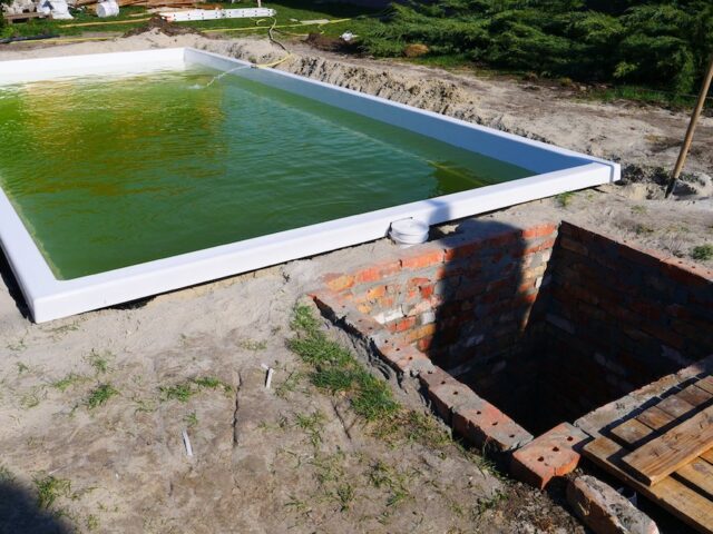 Белая чаша бассейна Эклипс, наполняющаяся водой в котловане рядом с сооружением кирпичного технического помещения.